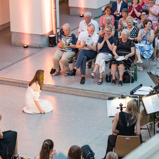 Eine Geigerin tritt in einem Konzertsaal auf, das Publikum sitzt im Hintergrund. Eine junge Frau in einem weißen Kleid sitzt auf der Bühne, und das Publikum schaut aufmerksam zu.