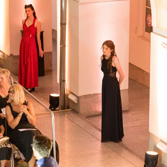 Eine Frau in einem roten Kleid steht in der Nähe einer Wand mit einem Spiegel, während eine andere Frau in Schwarz zusieht. Der Raum hat eine rosa Wand, ein Metallgeländer und zwei Lampen.