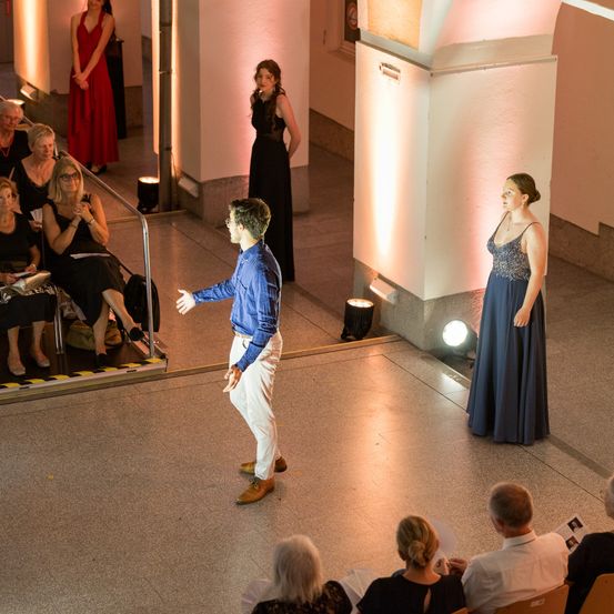Ein Mann in einem blauen Hemd und weißen Hosen spricht zu einer Frau in einem blauen Kleid in einem schwach beleuchteten Raum. Mehrere Leute sitzen im Hintergrund, darunter eine Frau in einem roten Kleid.