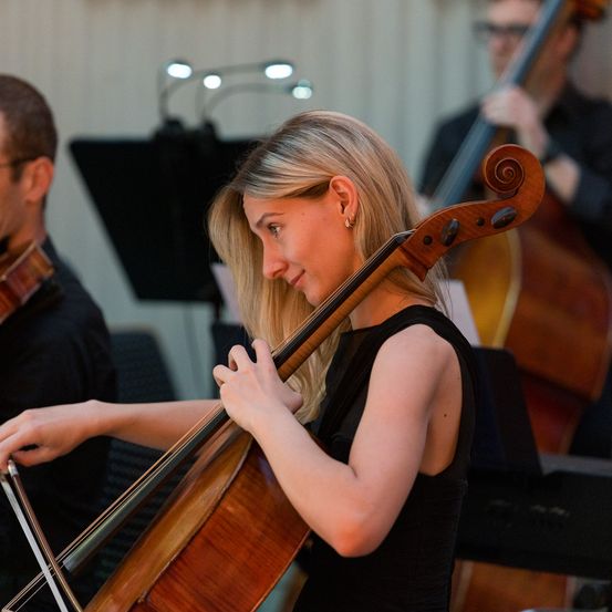 Eine Frau spielt Cello und lächelt, mit einem Geiger hinter ihr. Sie sind in einem Studio, mit einem Cellisten im Hintergrund.