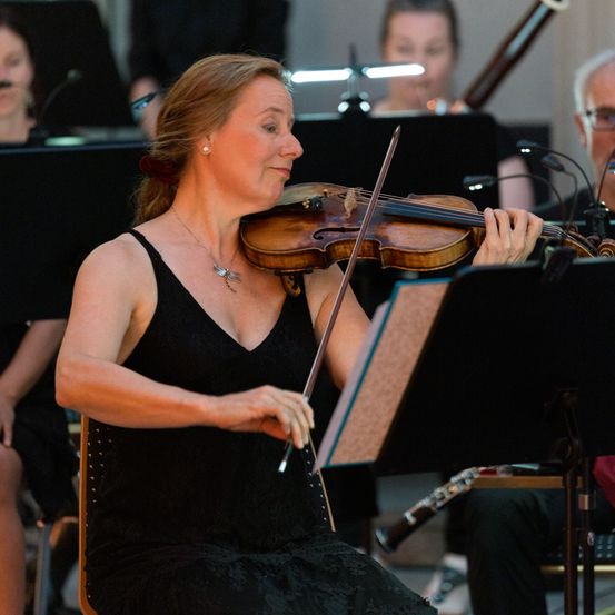 Eine Frau spielt eine Geige während einer musikalischen Aufführung, mit einer Band hinter ihr, einige Musiker spielen ihre Instrumente.