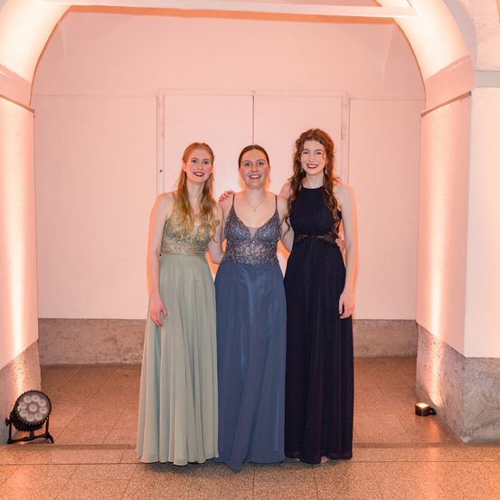Drei Frauen in Abendkleidern stehen eng zusammen in einem Flur. Sie lächeln und scheinen für ein Foto zu posieren.