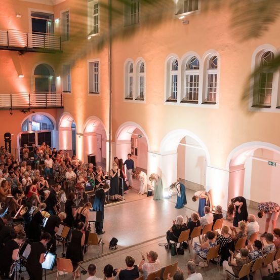 Eine große Gruppe von Menschen versammelt sich in einem Innenhof, in dem ein Orchester vor ihnen spielt. Der Innenhof hat Rundbogenfenster und ist mit warmer Beleuchtung erleuchtet. Einige Leute sitzen auf Stühlen, während andere stehen.