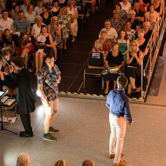 Zwei Männer stehen auf der Bühne, einer hält eine Taschenlampe. Ein Violinist spielt im Hintergrund. Viele Menschen sitzen auf Stühlen und schauen zu.