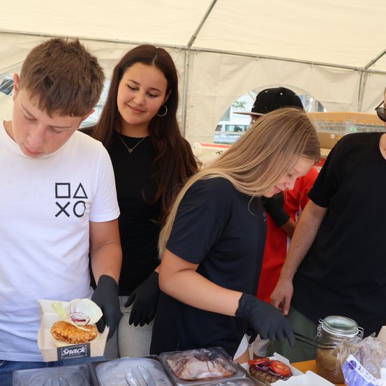 Vier Personen in einem Zelt bereiten Essen zu. Ein Mann in einem weißen T-Shirt hält einen Burger, eine Frau in Schwarz beugt sich über eine Schale und zwei weitere stehen dahinter.