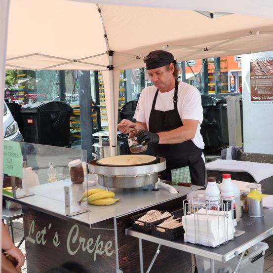 Ein Mann in einem weißen T-Shirt und einer schwarzen Schürze kocht Crepes auf einem Crepe-Stand mit Bananen und Gewürzen auf einem Tisch.