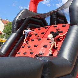 Zwei Kinder klettern auf einer roten aufblasbaren Kletterwand, eines in einem blauen Shirt und das andere in Schwarz. Die Wand ist schwarz und rot mit einem blauen Himmel im Hintergrund.