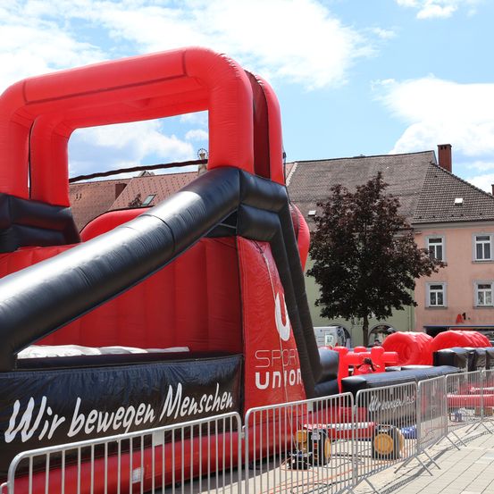 Ein großer roter und schwarzer aufblasbarer Hindernisparcours mit den Worten 'Wir bewegen Menschen' steht auf einem Stadtplatz. Dahinter befindet sich ein Gebäude mit Schornstein und einem Baum.