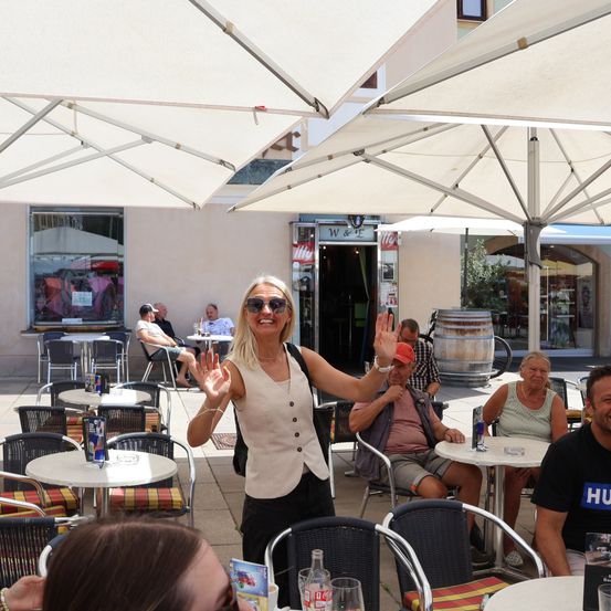 Eine Frau steht in einem Café im Freien und winkt zur Kamera, während mehrere Personen unter Sonnenschirmen an Tischen sitzen.