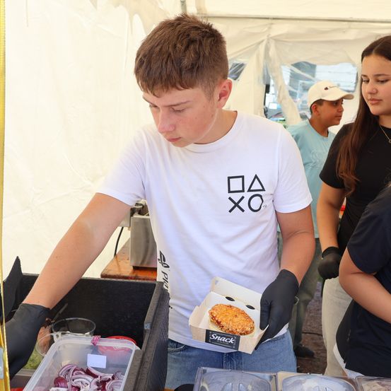 Ein junger Mann in einem weißen T-Shirt mit PlayStation-Logo hält eine Snackbox in einem Zelt. Hinter ihm stehen ein Mädchen und ein Junge.