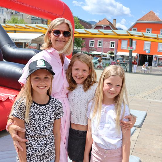 Eine Frau und drei junge Mädchen stehen vor einer bunten aufblasbaren Struktur auf einem Stadtplatz. Die Frau trägt eine Sonnenbrille und ein rosa Hemd. Die Mädchen tragen Freizeitkleidung und lächeln für die Kamera.