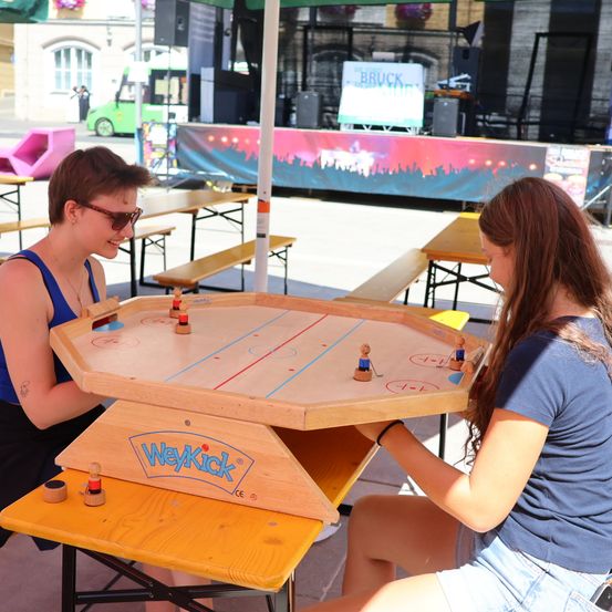 Zwei Frauen spielen Tischhockey im Freien. Das Spiel ist auf einem Holztisch mit einem Eishockeyfeld-Design aufgebaut. Die Frauen sitzen unter einem Schirm. Im Hintergrund befindet sich eine Bühne mit einem Banner und einem Bus.