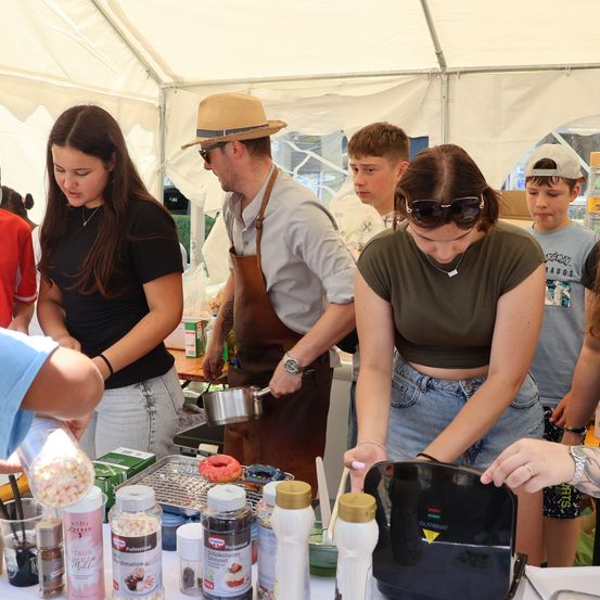 Eine Gruppe von Menschen versammelt sich in einem Zelt um einen Tisch und bereitet Essen zu. Ein Mann in einem Hut kocht, während eine Frau mit Sonnenbrille einen Nachtisch dekoriert. Verschiedene Zutaten und Utensilien liegen auf dem Tisch.