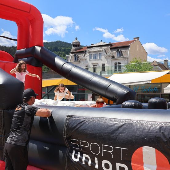 Eine Gruppe von Kindern spielt in einem großen aufblasbaren Hindernisparcours, den ein Mann beaufsichtigt. Der Aufblasbare ist mit dem Wort Sport Union gekennzeichnet. Im Hintergrund befinden sich Gebäude und ein Berg.