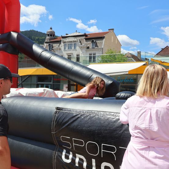 Ein junges Mädchen rutscht auf einem schwarzen aufblasbaren 'SPORT UNION'-Rutschvergnügen, während ein Mann und eine Frau zusehen. Gebäude und Berge sind im Hintergrund.