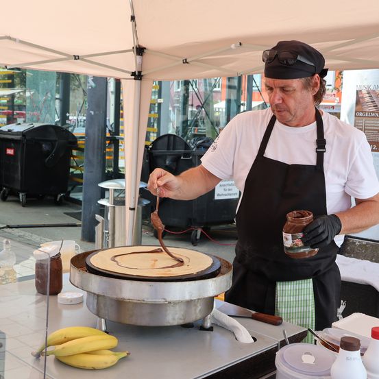 Ein Mann in einem schwarzen Kochschürze und Hut bereitet einen Crêpe mit Schokoladensauce auf einem Crêpe-Maker zu. Er hält eine Dose Schokoladensauce und einen Löffel. Bananen und andere Zutaten sind auf dem Tisch.