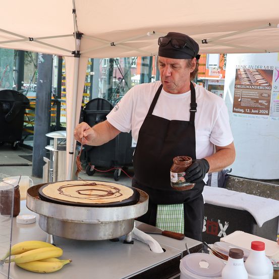 Ein Mann in einer schwarzen Schürze bereitet einen Crêpe mit Schokoladensauce auf einem Crêpe-Maker unter einem Zelt zu.