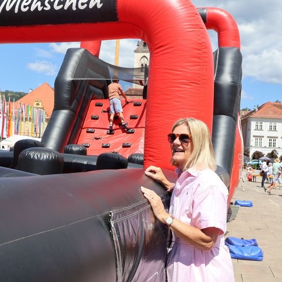 Eine Frau steht neben einem großen aufblasbaren Hindernisparcours, auf dem ein Junge klettert. Die Szene ist im Freien mit Gebäuden und einem Turm im Hintergrund.