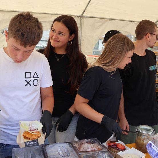 Eine Gruppe von Menschen in einem Zelt bereitet Essen zu, einer hält einen Burger, andere schauen in Essensbehälter.