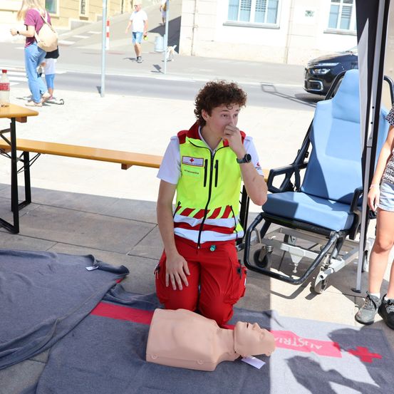 Ein junger Mann in einer gelben Sicherheitsweste übt CPR an einem Dummies auf dem Bürgersteig. Eine Frau in einem Blumenrock steht in der Nähe.