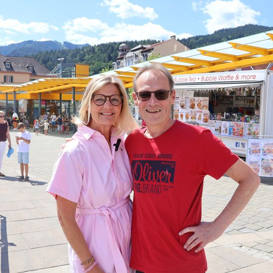 Ein Mann und eine Frau stehen lächelnd für ein Foto auf einem Stadtplatz. Hinter ihnen befindet sich ein Imbisswagen mit gelbem Vordach.