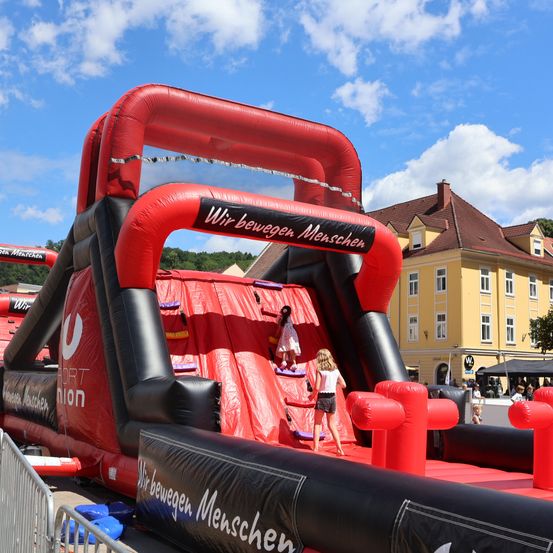 Zwei Kinder klettern auf einem roten aufblasbaren Hindernisparcours mit der Aufschrift 'Wir bewegen Menschen', vor dem Hintergrund eines gelben Gebäudes mit braunem Dach und einem blauen Himmel mit Wolken.