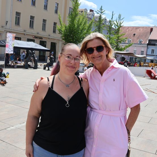 Zwei Frauen stehen in einem Außenbereich, eine trägt ein schwarzes Tanktop und die andere ein rosa Hemd. Sie lächeln und posieren für ein Foto. Hinter ihnen befinden sich Gebäude, Bäume und Menschen.
