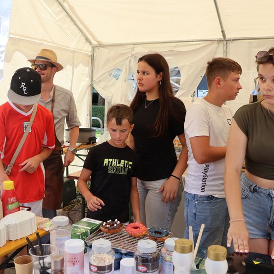 Eine Gruppe junger Leute steht um einen Tisch mit Donuts und Toppings. Ein Junge in einem schwarzen T-Shirt mit 'Athletic Skills' drauf schaut sich die Donuts an.