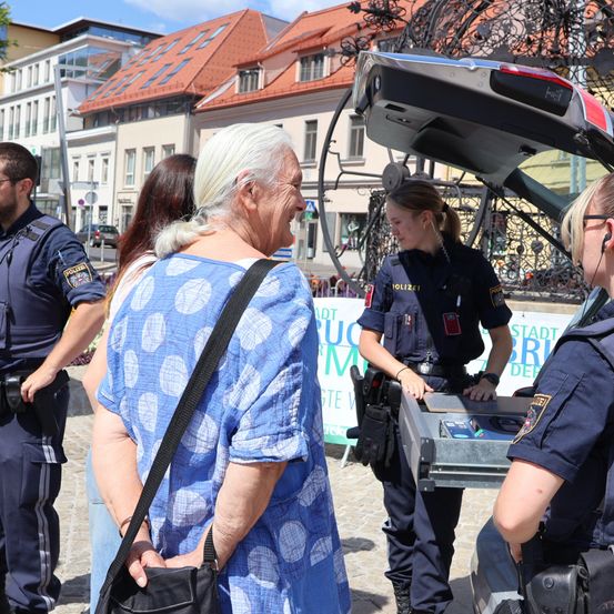 Eine Gruppe von Polizeibeamten interagiert mit einer älteren Frau auf einem Stadtplatz, mit Gebäuden und einem Auto im Hintergrund.