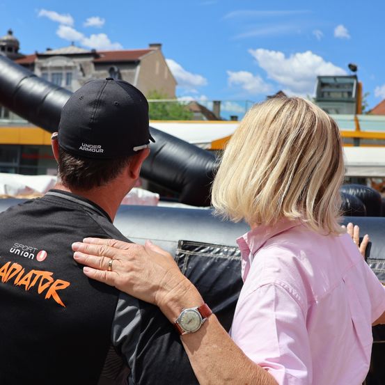 Ein Mann in einer schwarzen Under Armour-Kappe und -Shirt steht mit der Hand auf der Schulter einer Frau, beide schauen auf eine große aufblasbare Struktur im Hintergrund.