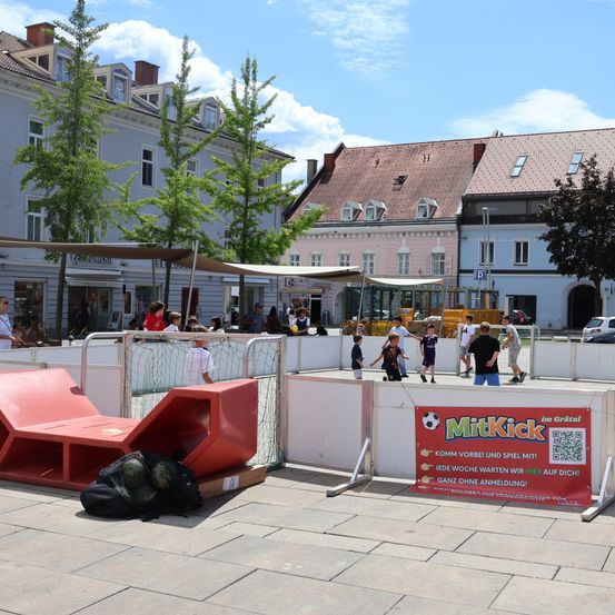 Eine Gruppe von Kindern spielt Fußball auf einem kleinen, eingezäunten Feld in einer Außenanlage. Das Feld ist von roten Barrieren umgeben und hat ein rotes Schild mit der Aufschrift 'MitKick'. Im Hintergrund befinden sich Gebäude und Bäume.