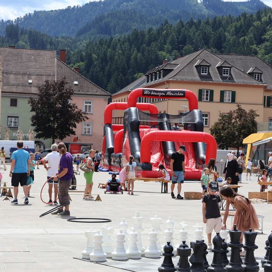 Eine Gruppe von Menschen versammelt sich auf einem Stadtplatz mit einem großen aufblasbaren Rutschen und einem riesigen Schachspiel auf dem Boden. Im Hintergrund befinden sich Gebäude und Bäume.
