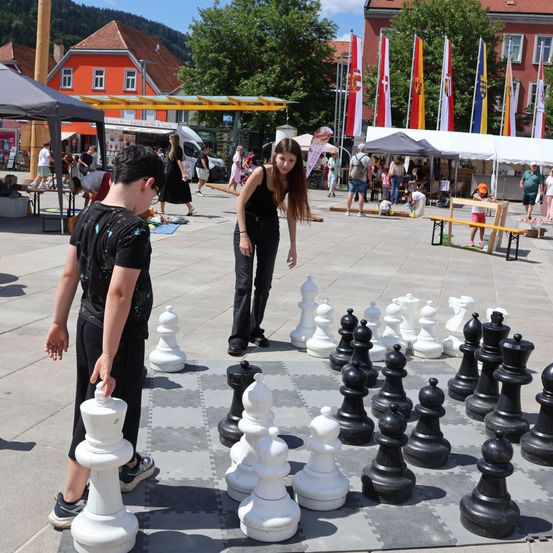 Zwei Personen spielen Schach auf einem großen Brett in einer Außenumgebung. Das Brett ist auf dem Boden aufgebaut, und die Figuren sind groß und weiß und schwarz.