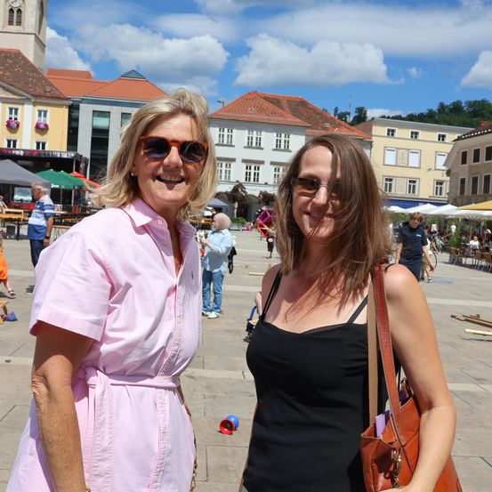 Zwei Frauen stehen auf einem Stadtplatz und lächeln für ein Foto. Hinter ihnen gehen und sitzen Leute unter Schirmen. Gebäude mit roten Dächern und weißen Fenstern sind sichtbar.