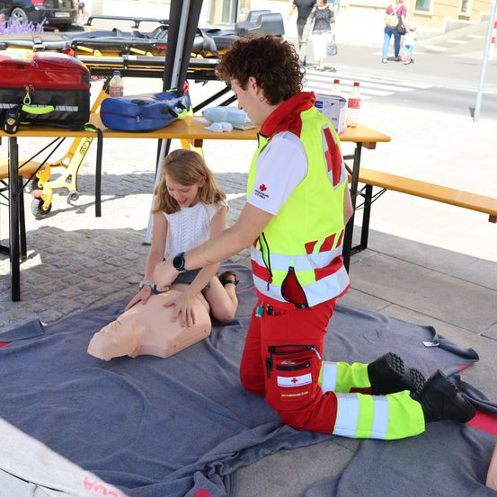 Ein Mann in einer Sicherheitsweste demonstriert einer kleinen Mädchen in einem weißen Kleid die Herzdruckmassage an einem Dummies. Sie befinden sich in einer Außenumgebung mit medizinischer Ausrüstung in der Nähe.