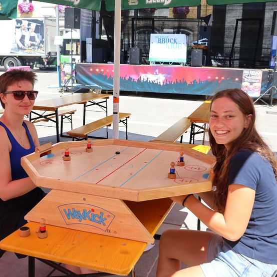 Zwei Frauen spielen Tischhockey bei einer Outdoor-Veranstaltung. Eine Frau trägt eine Sonnenbrille und eine Halskette, die andere trägt ein blaues Shirt und Shorts. Das Spiel ist auf einem Holztisch aufgebaut, über dem ein grüner Schirm steht. Im Hintergrund befindet sich eine Bühne mit einem Banner und einem Lastwagen.