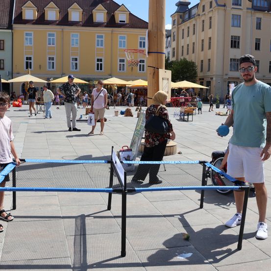 Eine Gruppe von Menschen spielt Tischtennis auf einem Stadtplatz, mit Gebäuden und Sonnenschirmen im Hintergrund.