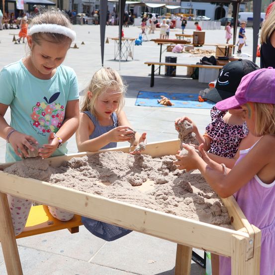 Vier junge Mädchen spielen in einem Sandkasten, jede hält ein kleines Objekt. Sie sitzen um einen mit Sand gefüllten Holztisch herum. Im Hintergrund gehen und stehen Leute, und einige sitzen auf Bänken.