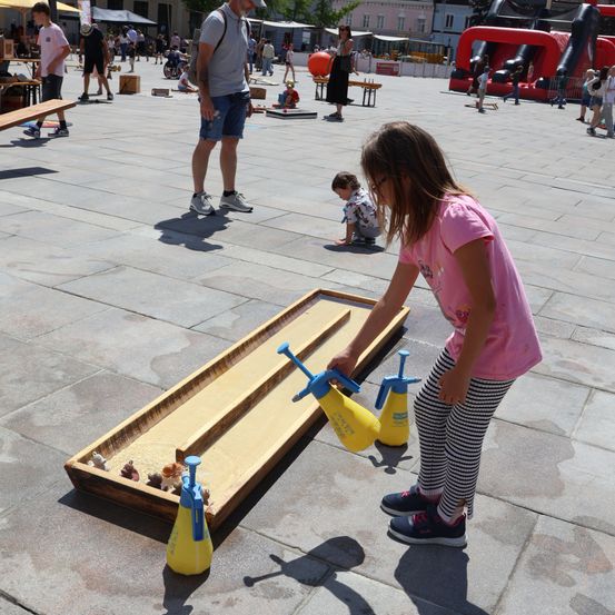 Ein junges Mädchen spielt ein Murmelspiel auf einer hölzernen Bahn auf einem öffentlichen Platz, mit anderen Kindern und Erwachsenen im Hintergrund.