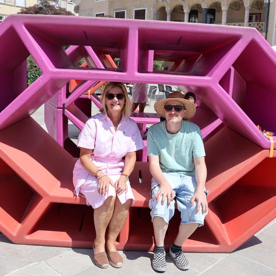 Eine Frau und ein Mann sitzen auf einer rosa geometrischen Bank im Freien. Die Frau trägt ein rosa Kleid und der Mann trägt ein grünes Shirt und einen Hut.