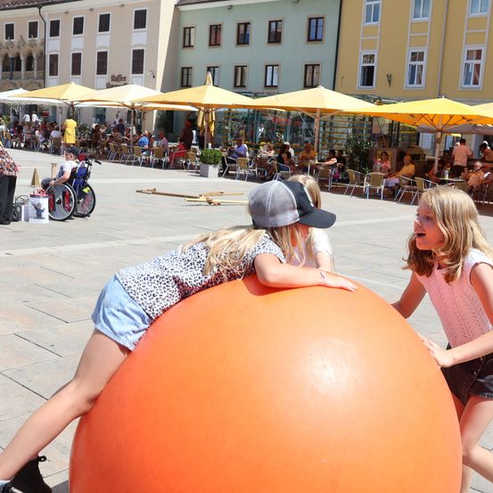 Zwei Mädchen spielen mit einem großen orangefarbenen Ball in einem Außenbereich mit Gebäuden und Menschen, die unter Sonnenschirmen an Tischen sitzen.
