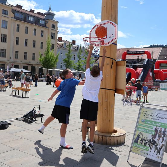 Zwei Jungen spielen Basketball an einem Holzpfosten auf einem Stadtplatz, mit einem Gebäude und aufblasbaren Attraktionen im Hintergrund.
