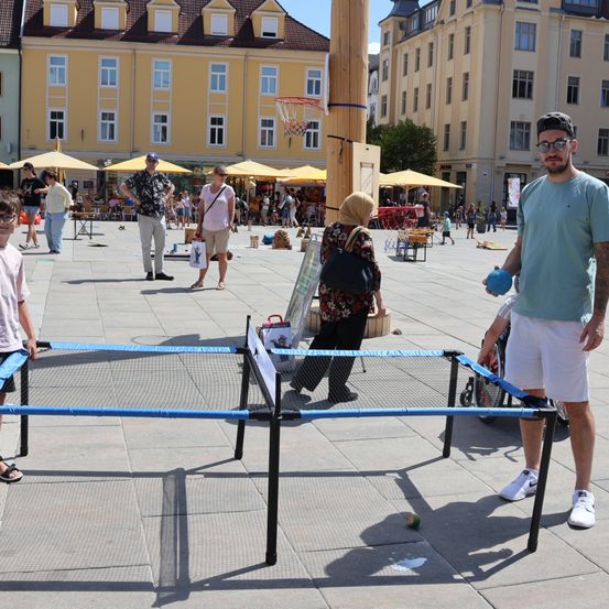 Eine Gruppe von Menschen spielt Tischtennis auf einem öffentlichen Platz mit Gebäuden und Schirmen im Hintergrund.
