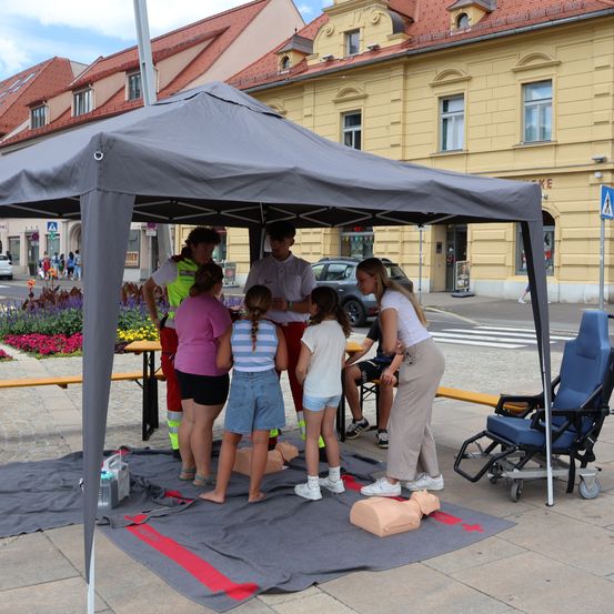 Eine Gruppe von Kindern und Erwachsenen versammelt sich unter einem Zelt und lernt Erste-Hilfe-Techniken von einem Mann in einer reflektierenden Weste.