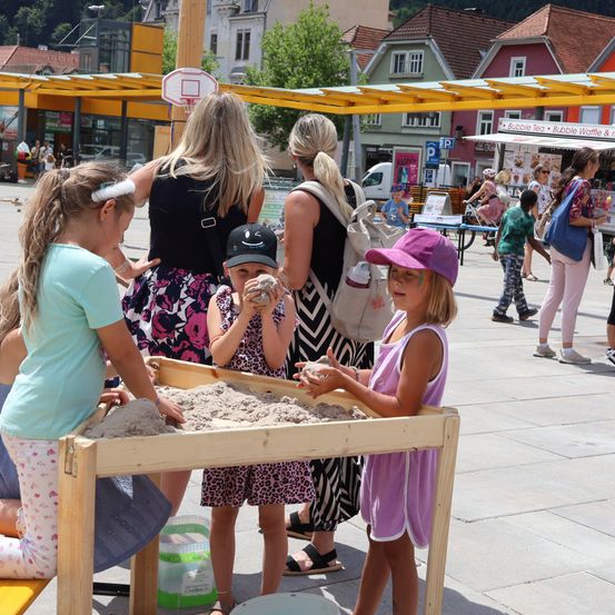 Vier junge Mädchen spielen in einem Sandkasten, ein Mädchen trägt einen lila Hut. Hinter ihnen trägt eine Frau einen Rucksack. Im Hintergrund befindet sich ein Gebäude mit einem Basketballkorb und einem Imbisswagen.
