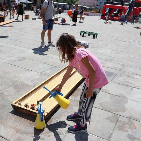 Ein junges Mädchen spielt ein wasserbasiertes Spiel auf einem Holzbrett in einem Außenbereich, während andere Menschen und aufblasbare Spielgeräte im Hintergrund zu sehen sind.