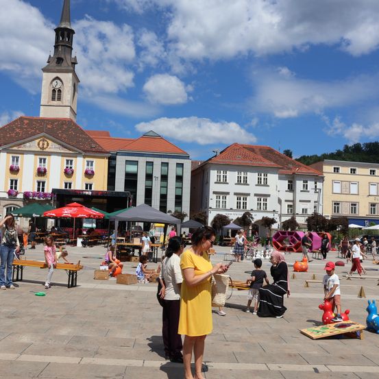 Ein sonniger Tag auf einem Stadtplatz mit einem Glockenturm, Menschen und spielenden Kindern auf dem Boden. Es gibt Bänke, Zelte und Spielzeug, die herum verstreut sind.