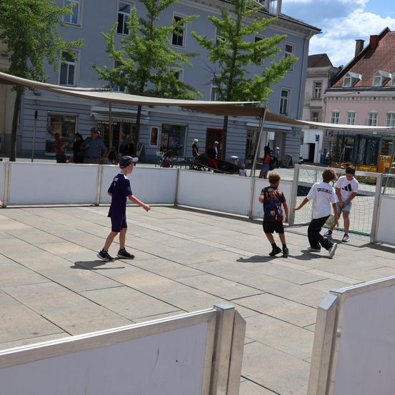 Vier Jungen spielen Fußball auf einem Betonplatz mit einer weißen Barriere, umgeben von Gebäuden und Bäumen.