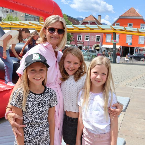 Eine Frau und drei Mädchen stehen vor einem leuchtend orangefarbenen Gebäude und lächeln für ein Foto. Dahinter sind ein Hüpfburg und ein Spielplatz zu sehen, mit Menschen und Autos im Hintergrund.