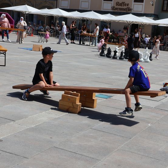 Zwei Jungen spielen Schach auf einer hölzernen Bank in einer geschäftigen Außenplatz, umgeben von Menschen und großen Sonnenschirmen.
